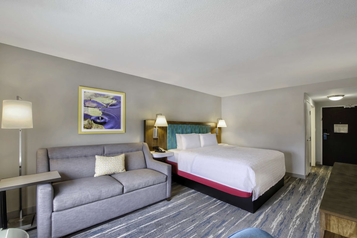 A hotel room with a grey sofa, a bed with white bedding, a nightstand with a lamp, and a painting on the wall.