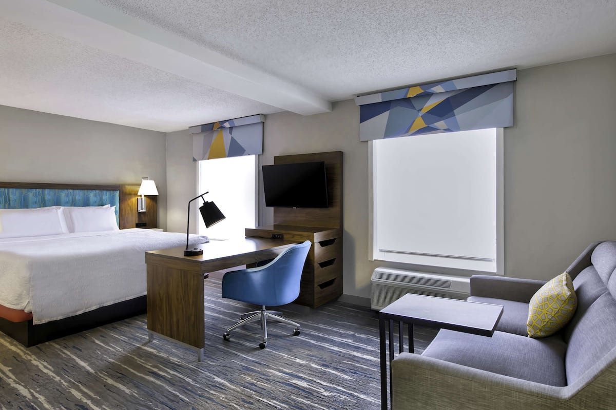 A hotel room with a bed, desk, chair, sofa, TV, and large windows.