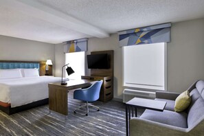 A hotel room with a bed, desk, chair, sofa, TV, and large windows.