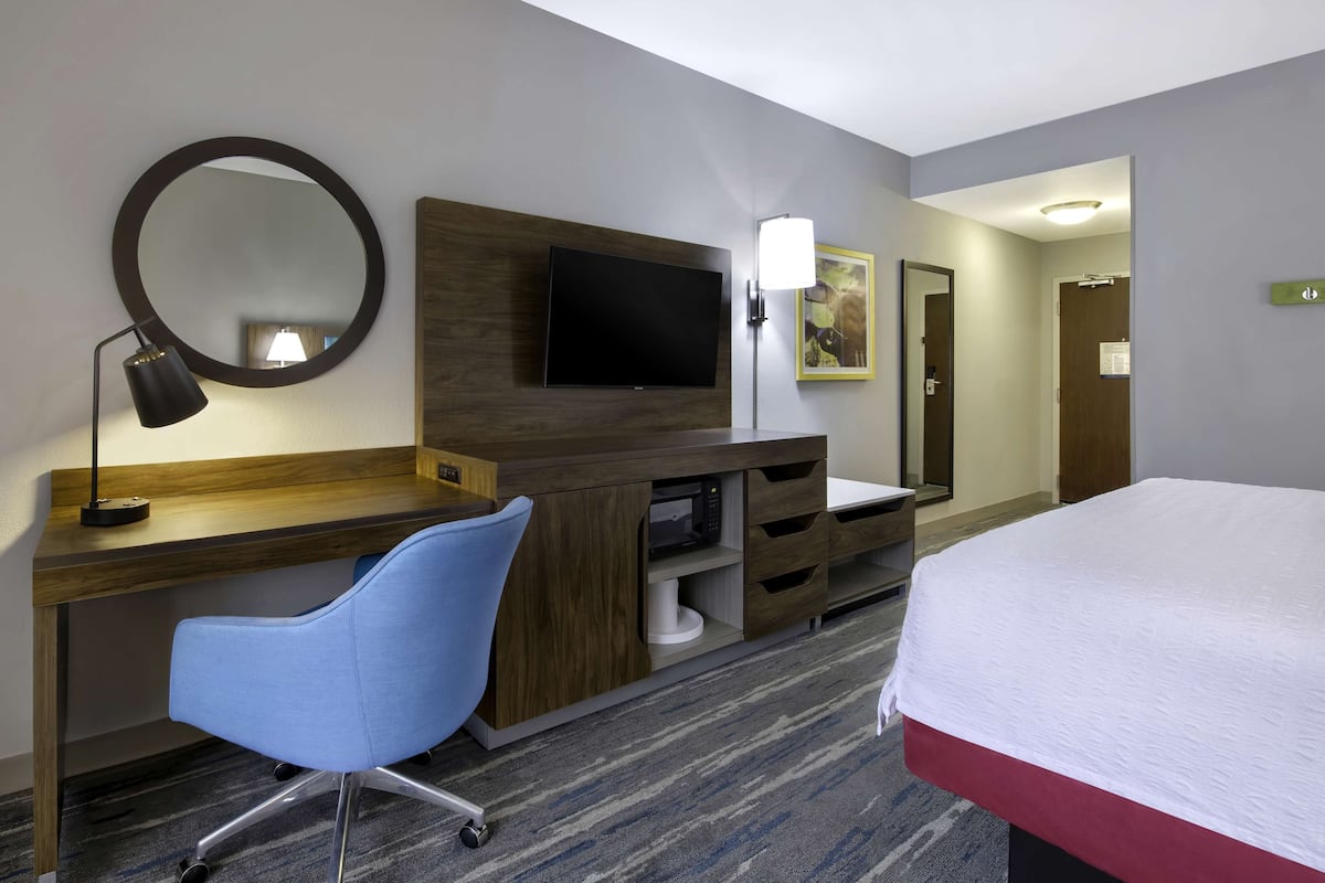 A hotel room with a bed, a desk with a chair, a TV, a mirror, and a lamp.