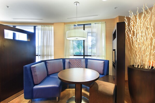 Una habitación de hotel moderna con un sofá seccional azul, una mesa redonda y un ventanal con cortinas.