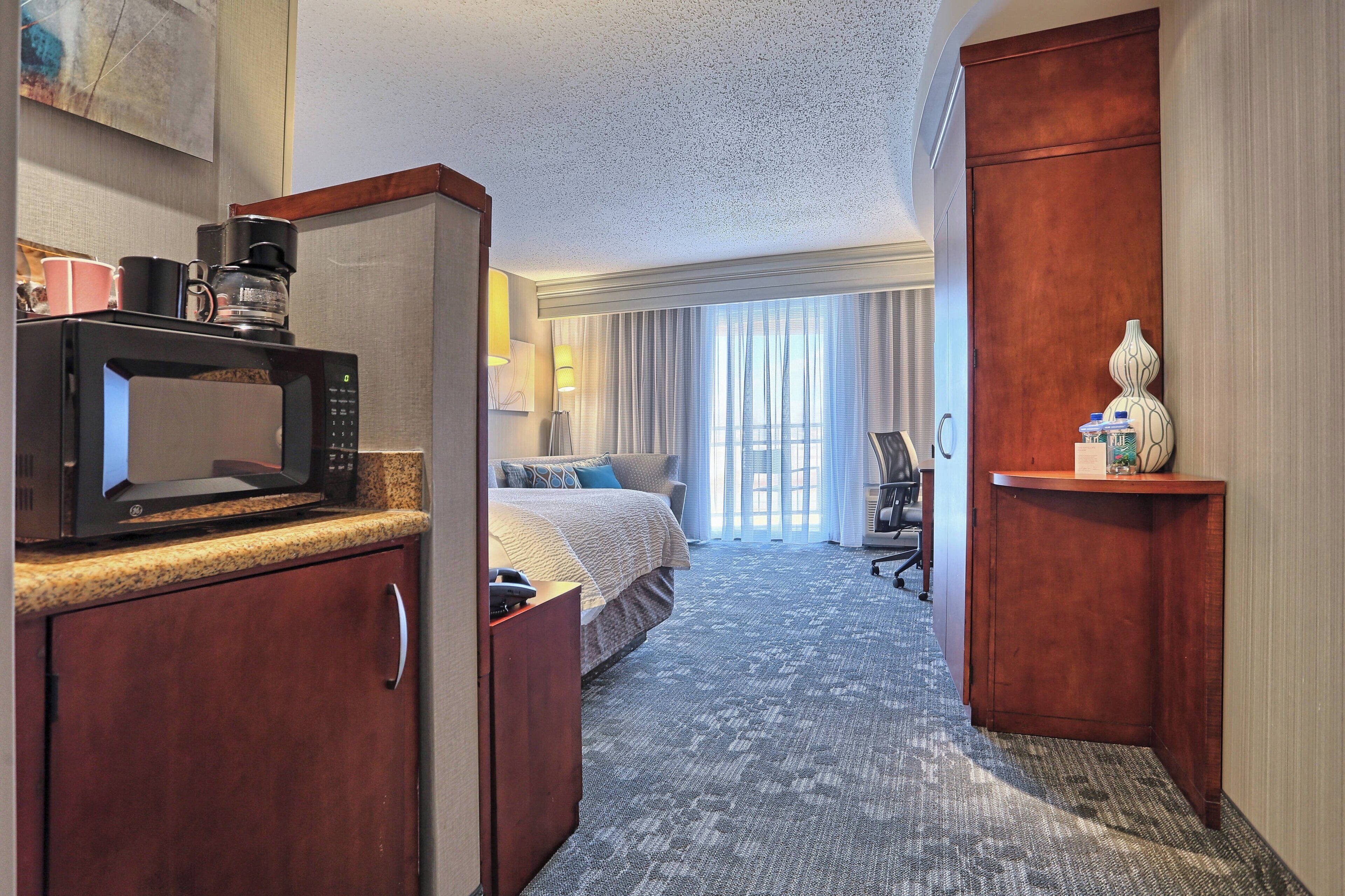 Habitación de hotel con kitchenette, cama sobre alfombra gris, escritorio con lámpara y ventana grande con cortinas.