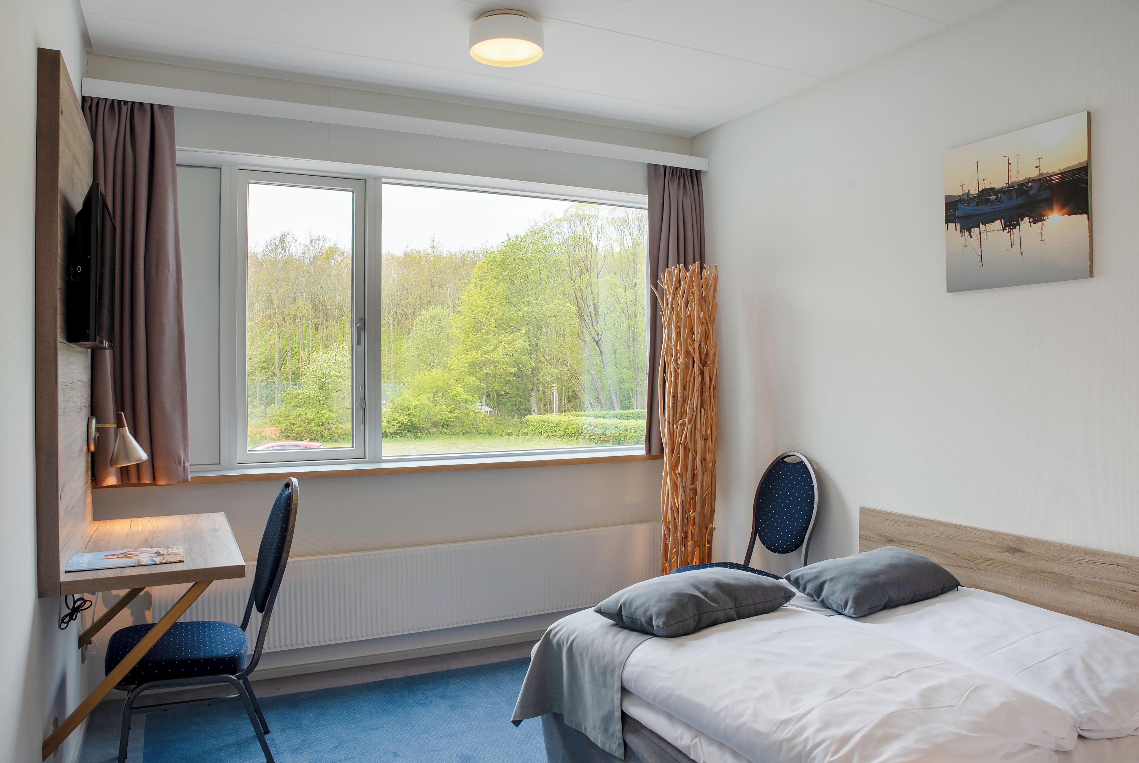 A bedroom with a bed, a desk, and a chair. There is a window with curtains, a radiator, and a painting on the wall.