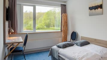 A bedroom with a bed, a desk, and a chair. There is a window with curtains, a radiator, and a painting on the wall.