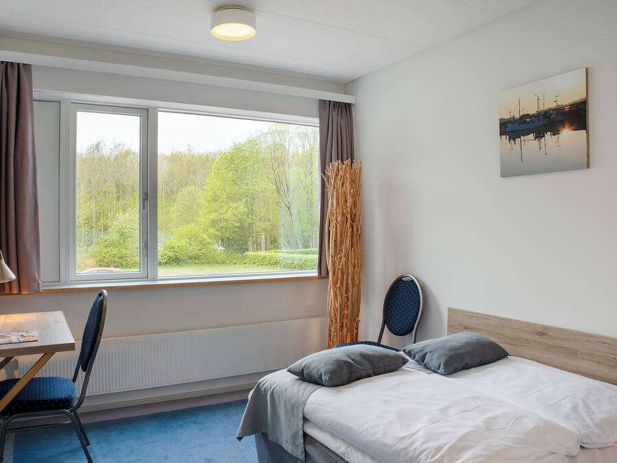 A bedroom with a bed, a desk, and a chair. There is a window with curtains, a radiator, and a painting on the wall.