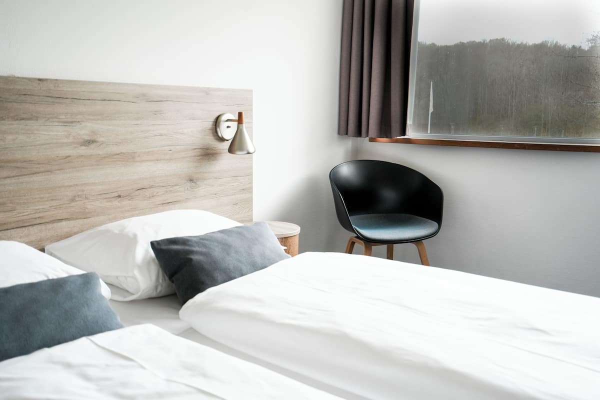 A modern hotel room with a wooden headboard, a black chair, and a large window with curtains.