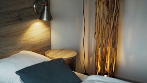 A neatly made bed with white linens and a dark pillow, a round wooden side table, a wall-mounted lamp, and a curtain with a natural, woven texture.
