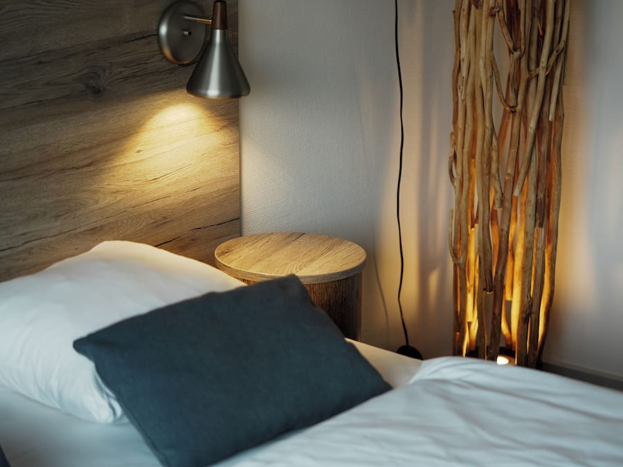 Una cama bien hecha con sábanas blancas y un almohadón oscuro, una mesita de madera redonda, una lámpara de pared y una cortina de textura natural y tejida.