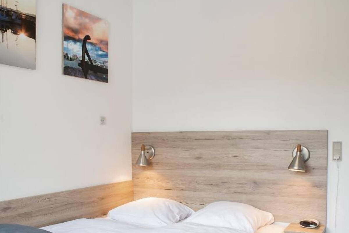A modern bedroom with a wooden headboard, two bedside lamps, a bed with grey and white bedding, and two wall-mounted artworks.