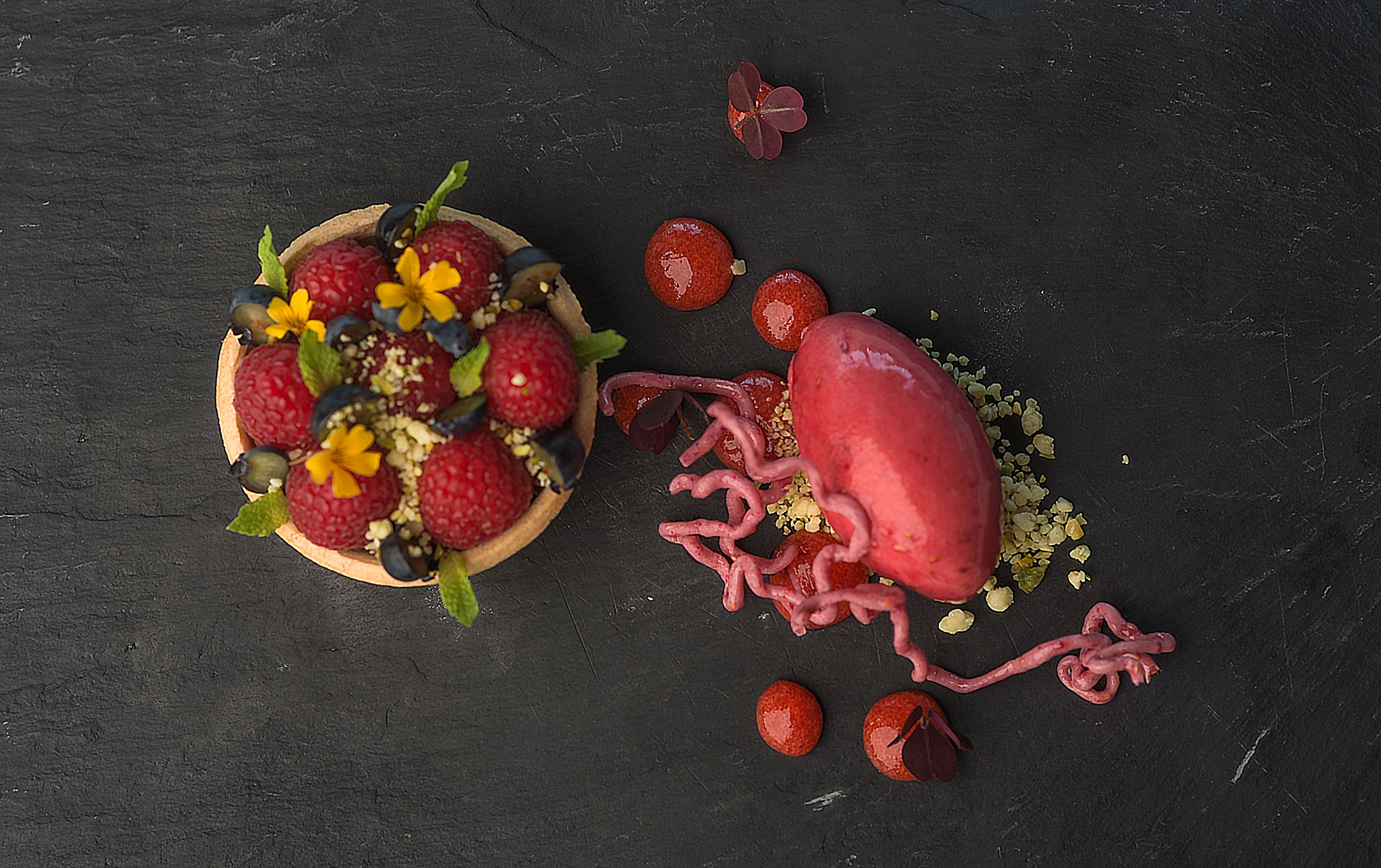En desserttallerken med hindbær, blåbær og en dekorativ blomst på en mørk overflade.
