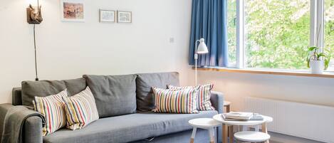 Una sala de estar moderna con un sofá gris, almohadones a rayas, una mesita redonda y un ventanal con cortinas azules.