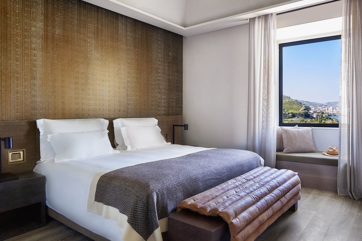 Ett modernt hotellrum med en stor säng, ett sängbord, ett fönster med utsikt över staden och en inbyggd bänk vid fönstret.