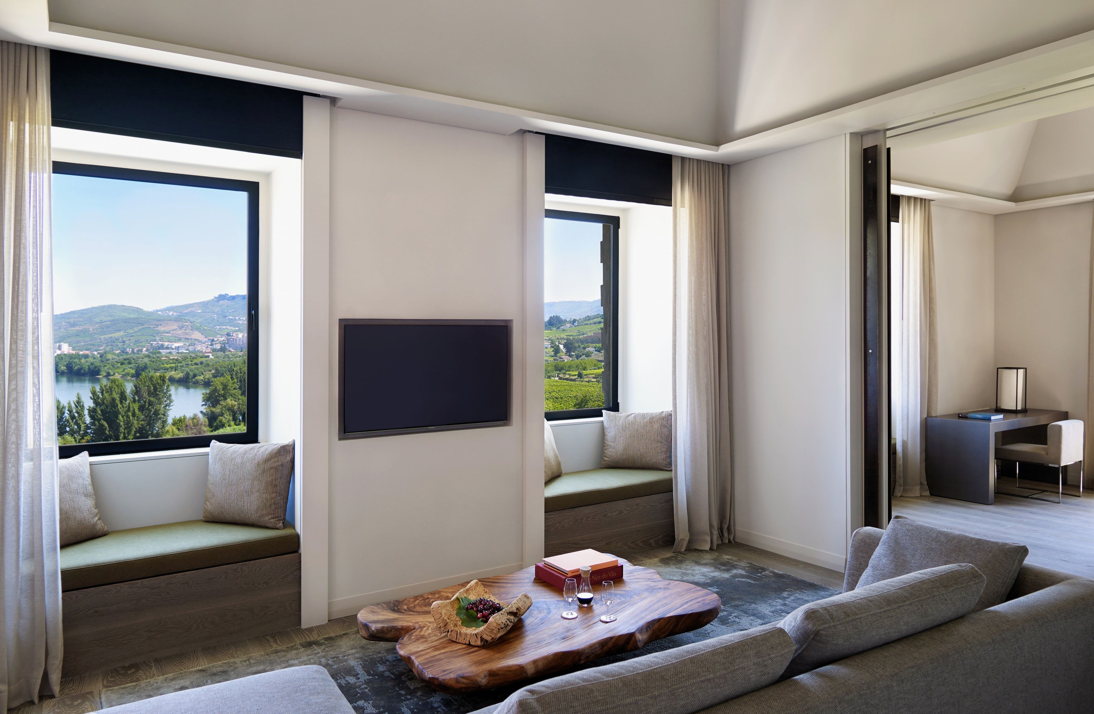 A modern hotel room with a large window offering a scenic view of a lake and hills. There is a cozy seating area with a sofa and a wooden coffee table with books and glasses.