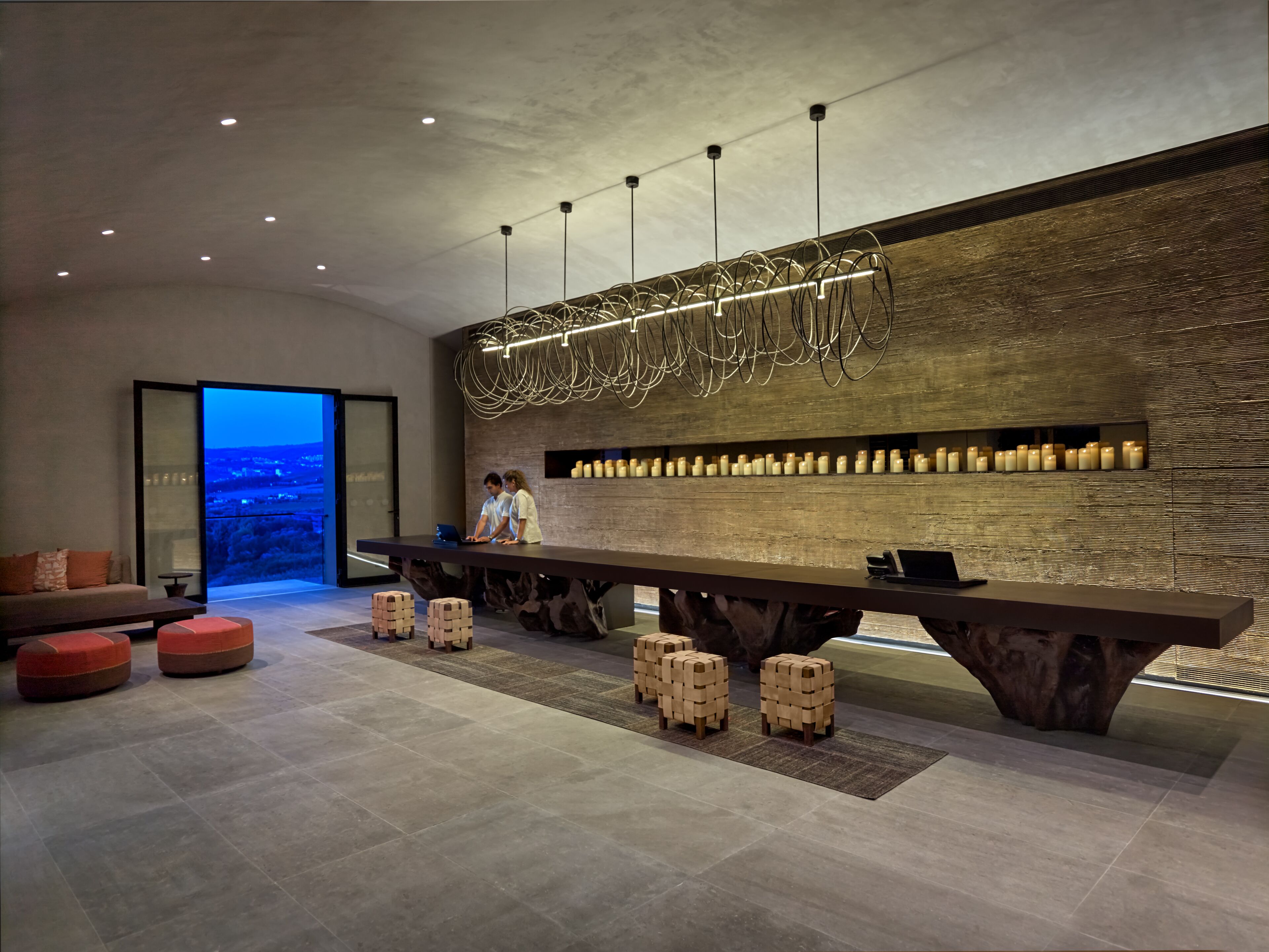 A modern interior with a long wooden table, red cushioned seating, and a view of a cityscape through a glass door.