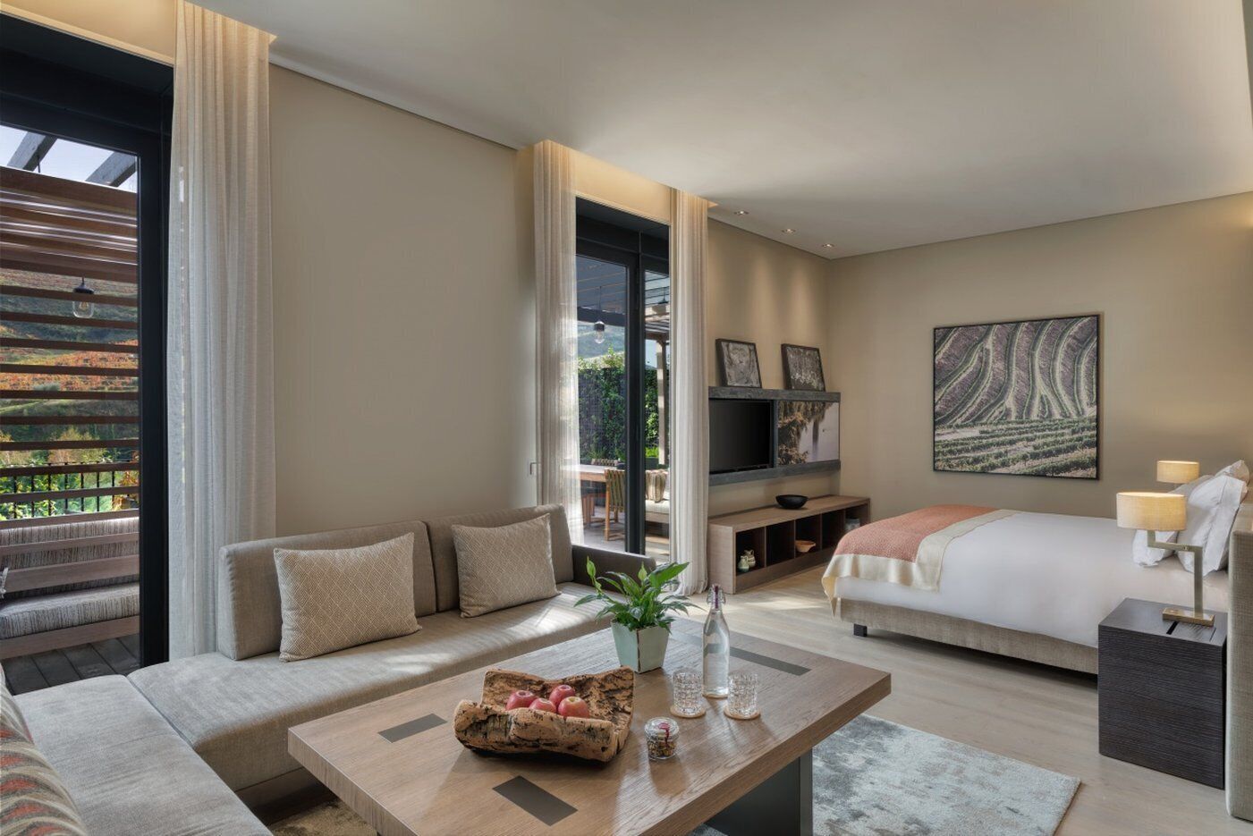 Habitación de hotel moderna con sofá, cama, televisor y zona de comedor con vistas al exterior.