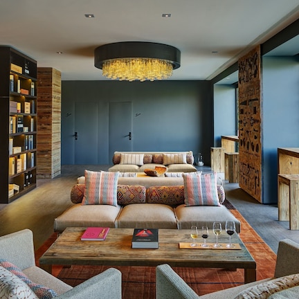 A modern living room with a sofa set, a coffee table with books and glasses, a bookshelf, and a large window with a view of greenery.