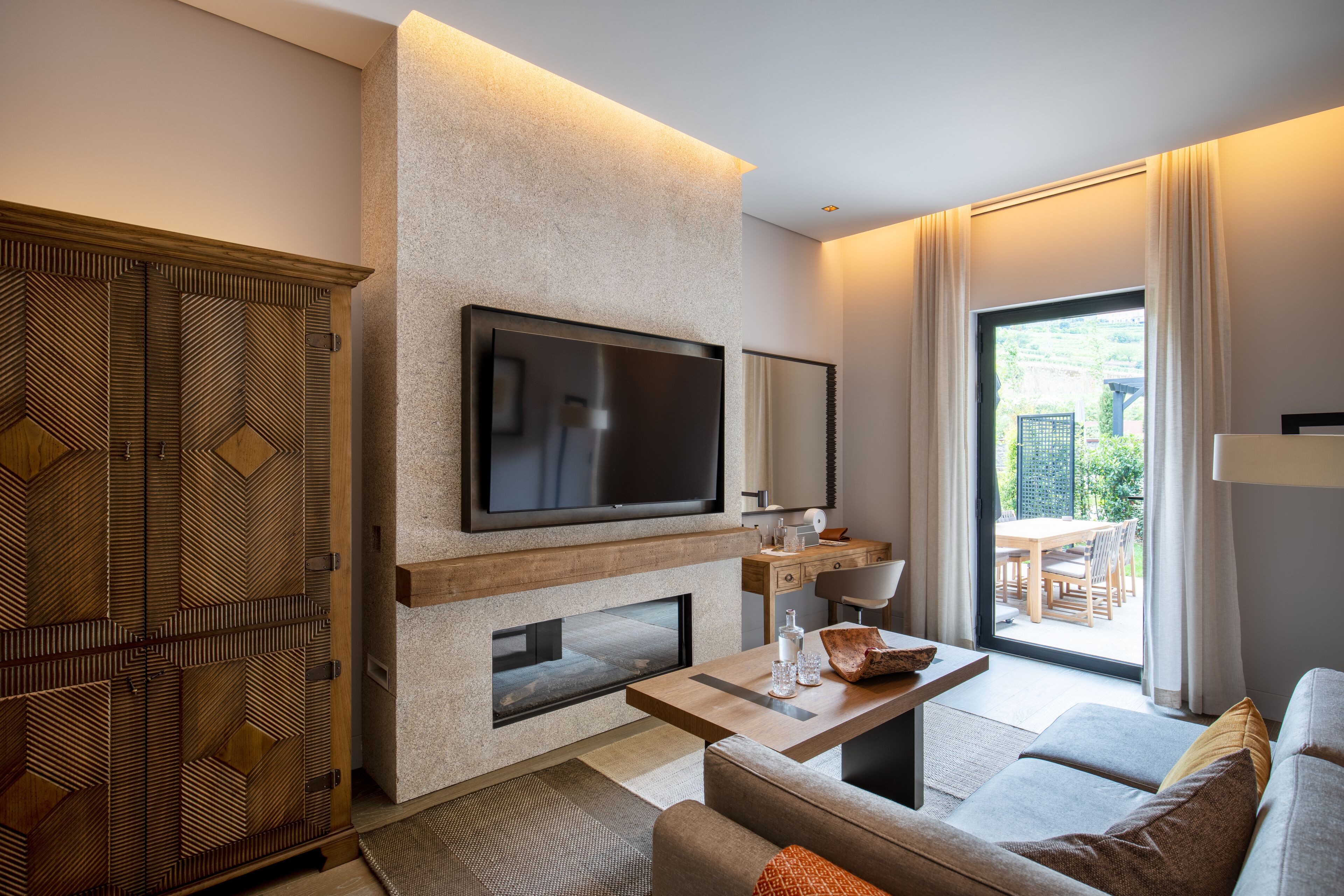 A modern living room with a flat-screen TV mounted on a textured wall, a fireplace, a sofa, and a dining area visible through a large window.
