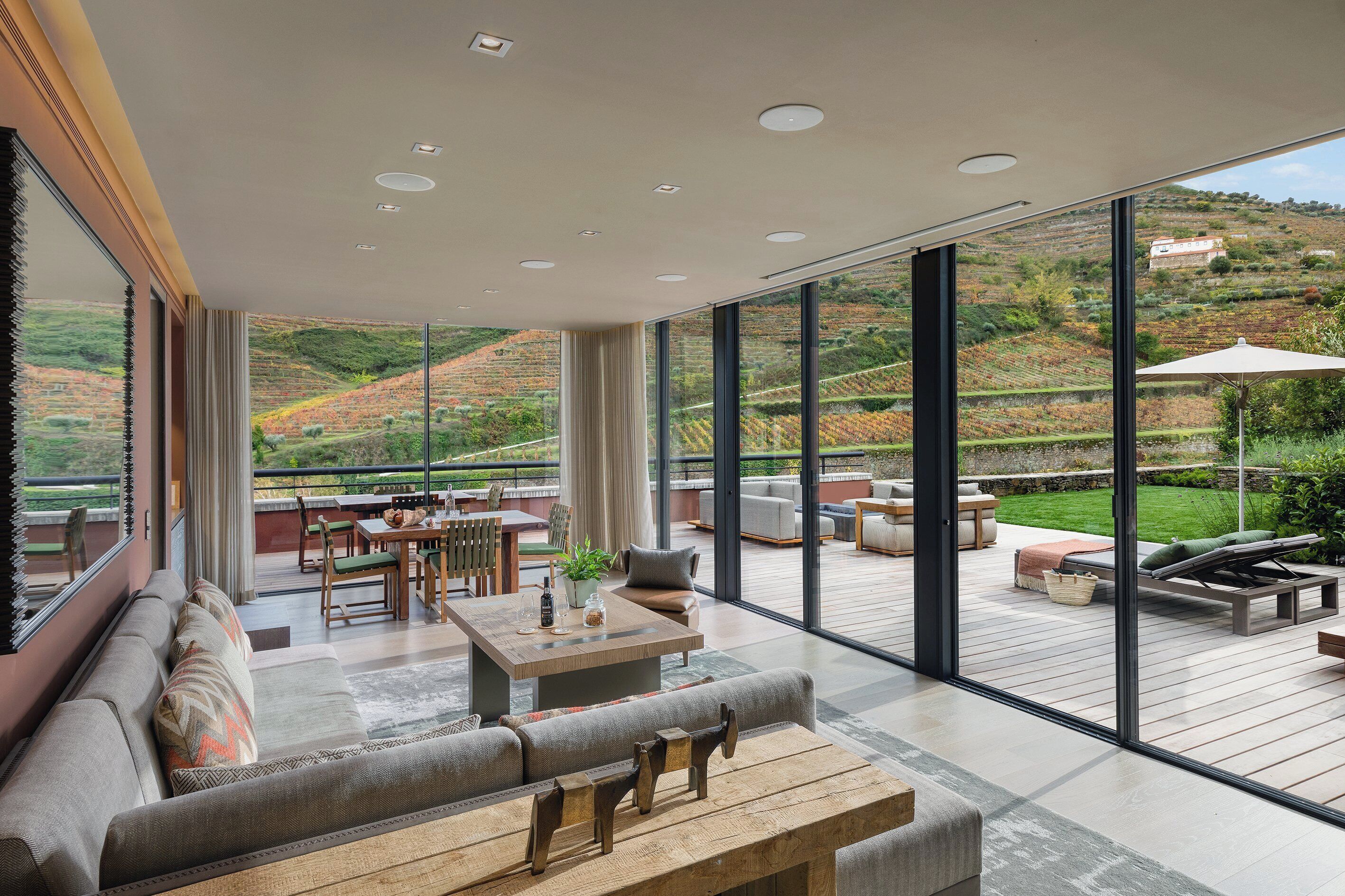 A spacious indoor area with a large window overlooking a vineyard. There is a wooden table with chairs, a sofa, and a lounge area with a view.