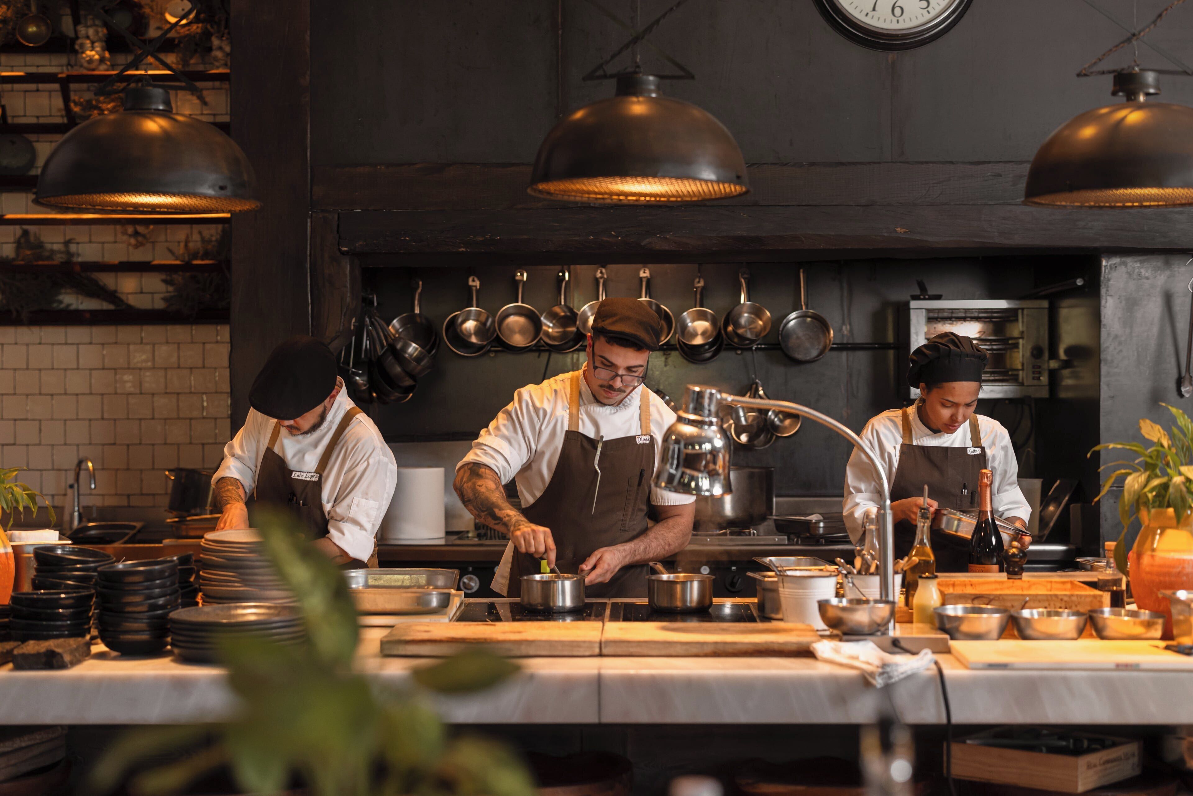 A professional kitchen with chefs preparing food, equipped with stainless steel appliances and hanging pots.