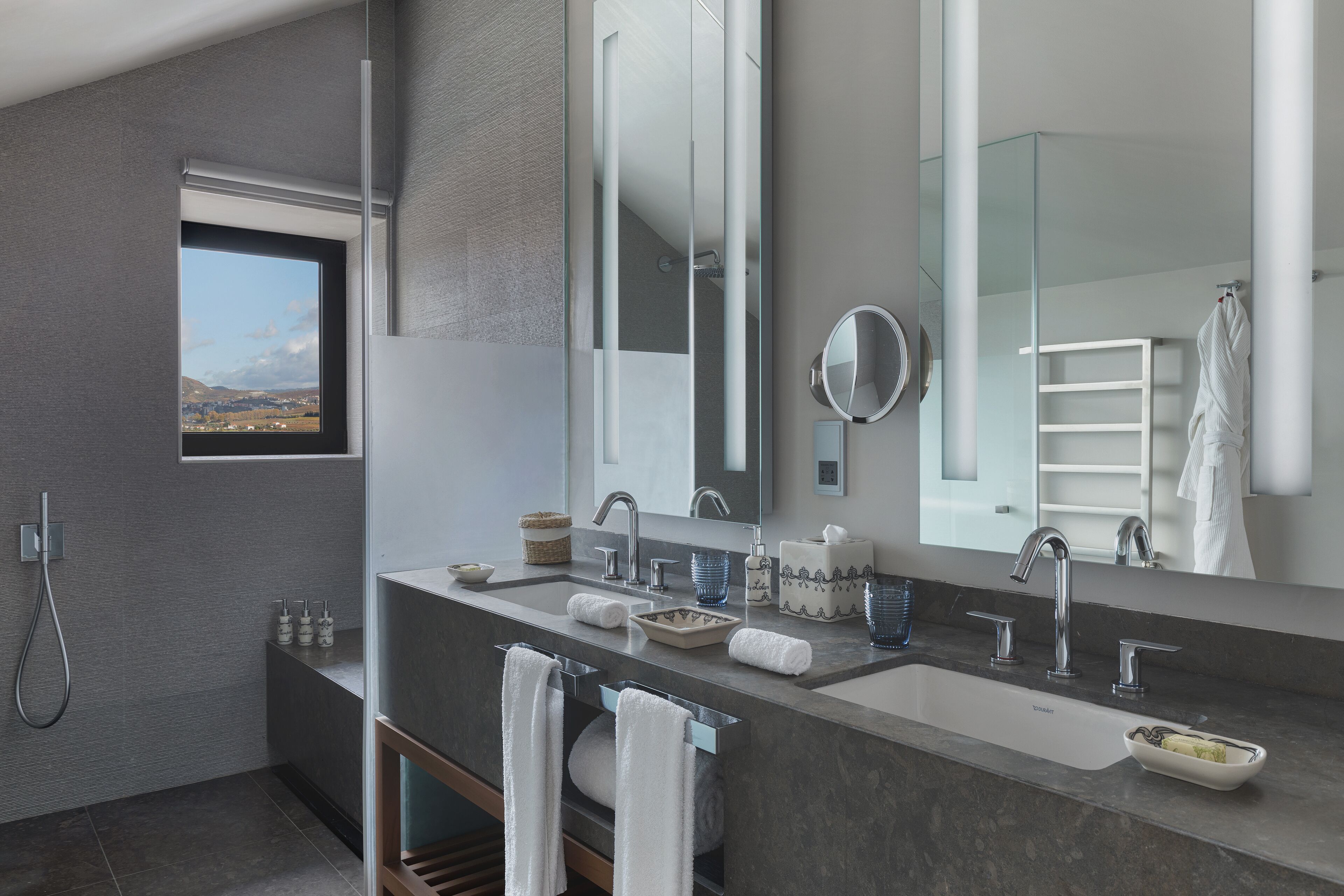 A modern bathroom with a large mirror, two sinks, a shower, and a window offering a view.