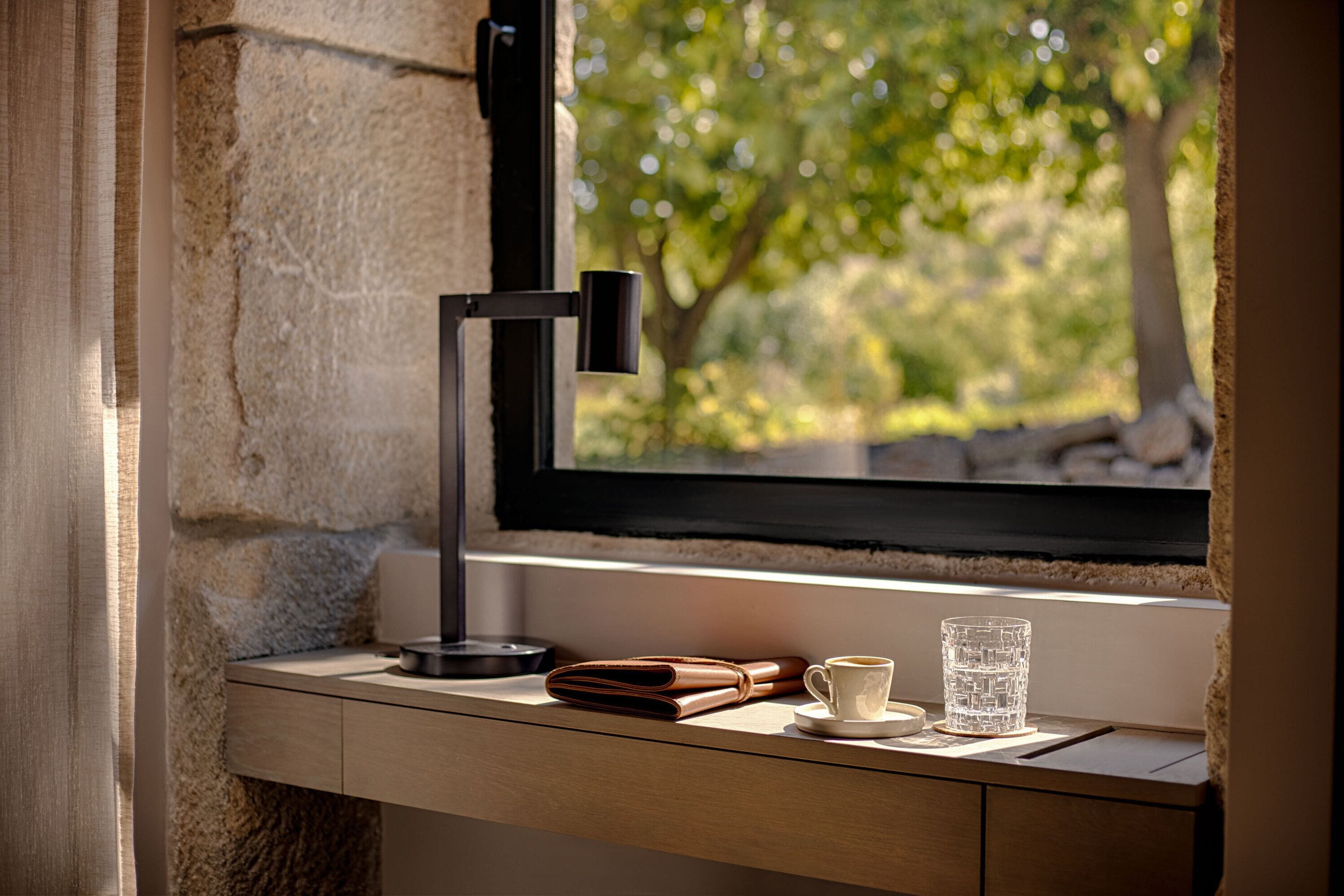 A desk with a laptop, a wallet, a cup, and a glass, positioned by a window with a view of trees.