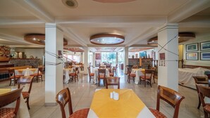 Un grand espace dédié à la salle à manger, avec plusieurs tables et chaises, une table centrale recouverte d’une nappe jaune, et une vue sur l’extérieur grâce à de grandes fenêtres.