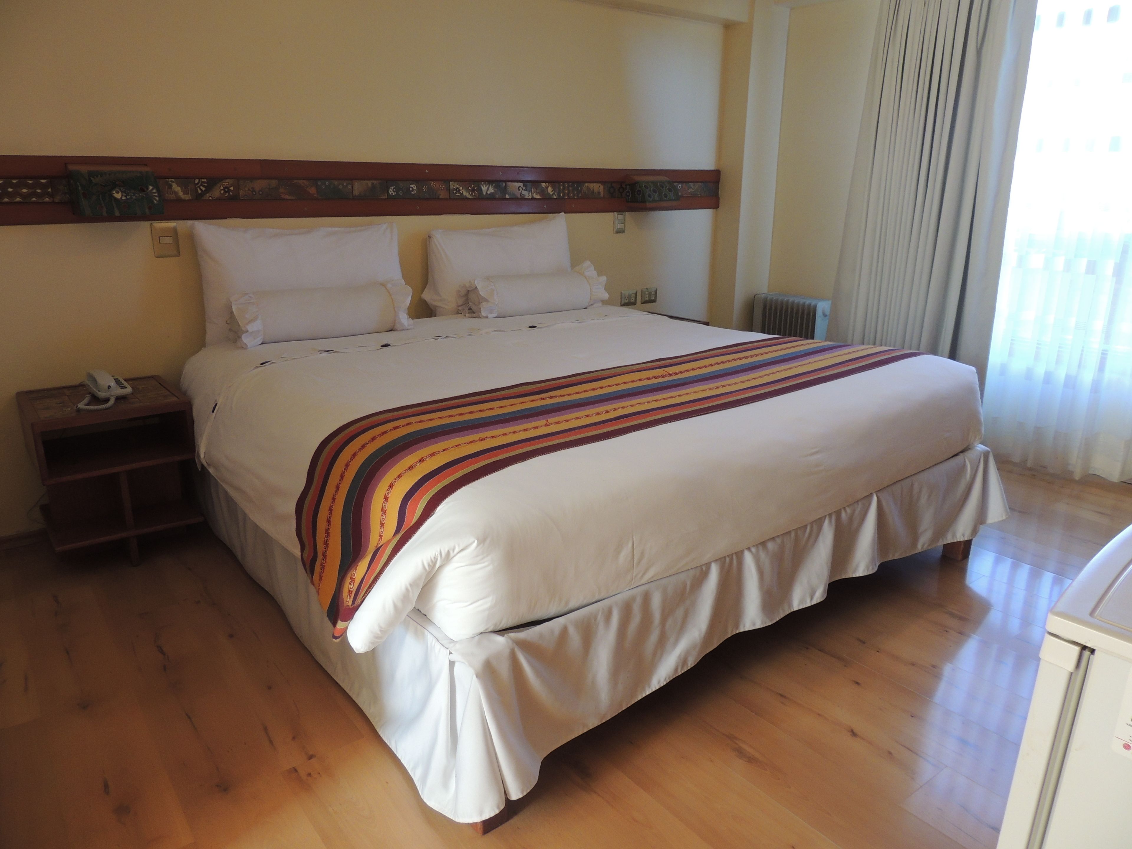 Una cama grande con una manta a rayas, cabecera de madera, mesita de noche con teléfono y ventana con cortinas.