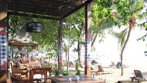 An outdoor seating area with wooden tables and chairs, a covered section with a sign reading "Batu-Bata Resort", and a beach with lounge chairs and umbrellas in the background. - Besakih Beach Hotel (Denpasar)