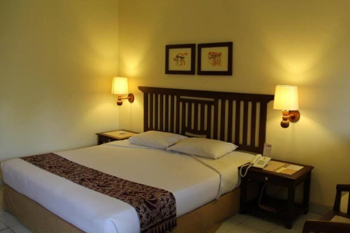 A hotel room with a bed, two bedside lamps, a nightstand with a phone, and two framed pictures on the wall.