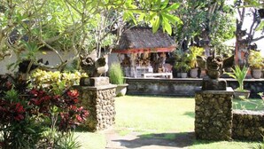 A garden with a stone pathway leading to a thatched-roof structure, surrounded by various plants and decorative statues. - Besakih Beach Hotel (Denpasar)