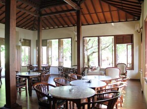 An indoor dining area with multiple round tables and chairs, large windows with shutters, and a wooden ceiling structure. - Besakih Beach Hotel (Denpasar)