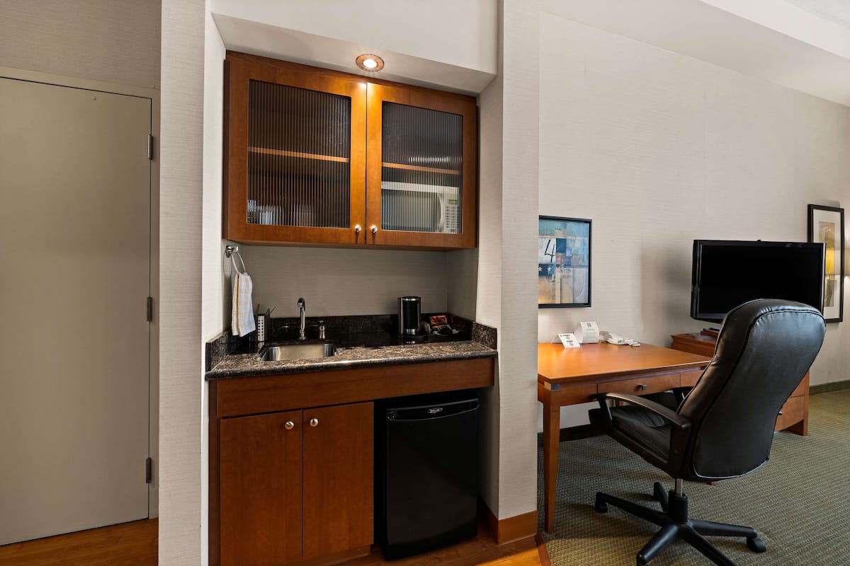 A compact hotel room with a kitchenette, a desk, a chair, and a television.