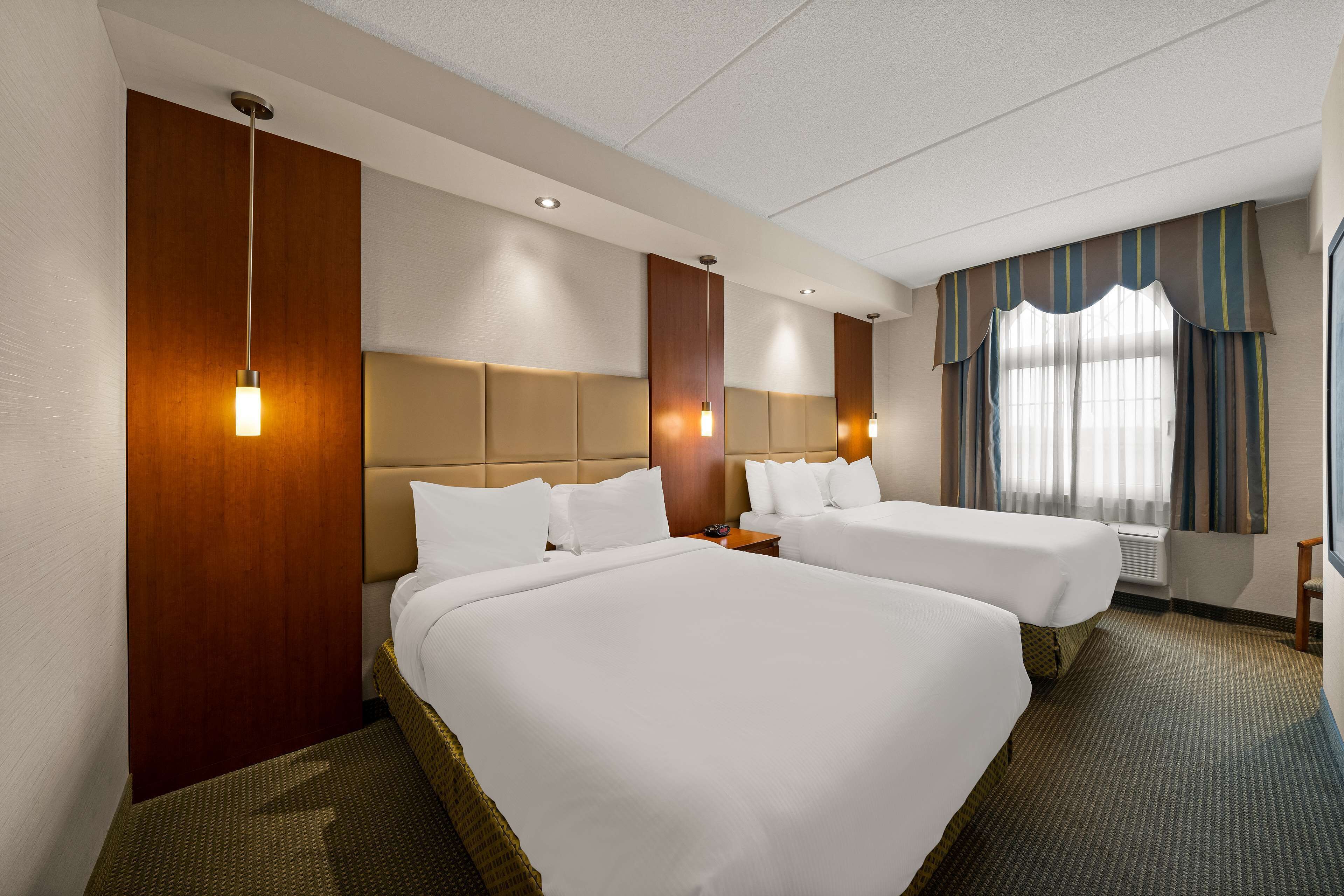 Habitación de hotel con dos camas, cabecero de madera, un ventanal grande con cortinas y una mesita pequeña.