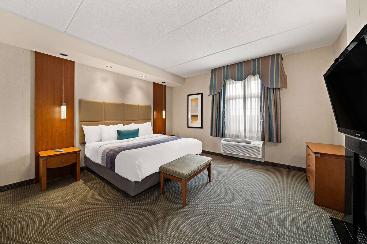 A hotel room with a bed, bedside table, bench, TV, and air conditioning unit.