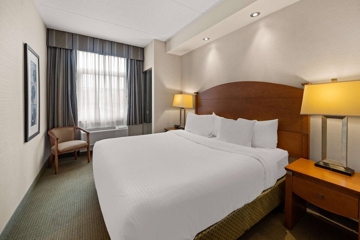A hotel room with a large bed, a wooden nightstand with a lamp, a chair, a window with curtains, and a framed picture on the wall.