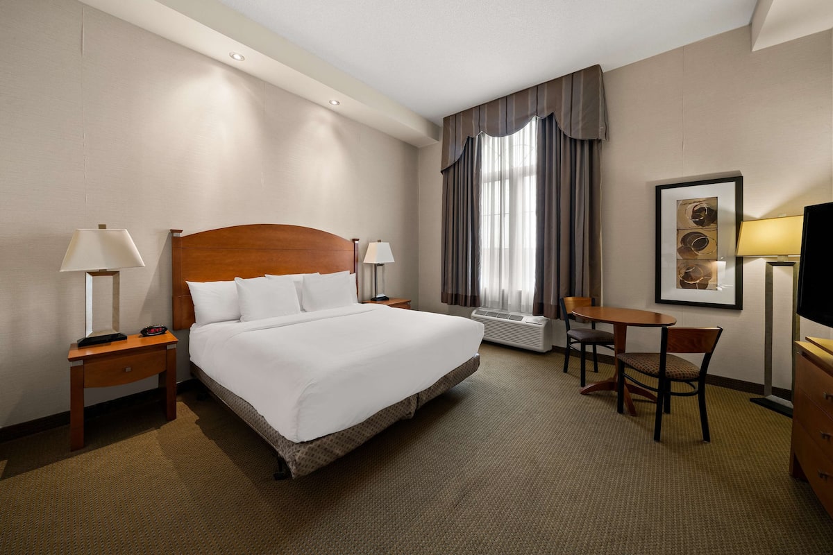 A hotel room with a large bed, two bedside tables with lamps, a desk with a chair, a television, and a framed picture on the wall.