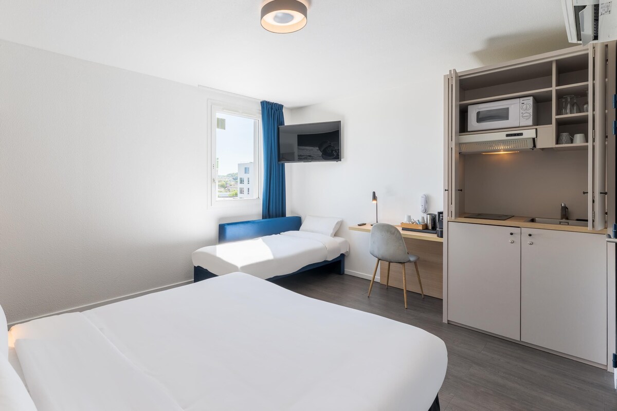 Una habitación de hotel con cama, escritorio, silla, televisor y kitchenette.