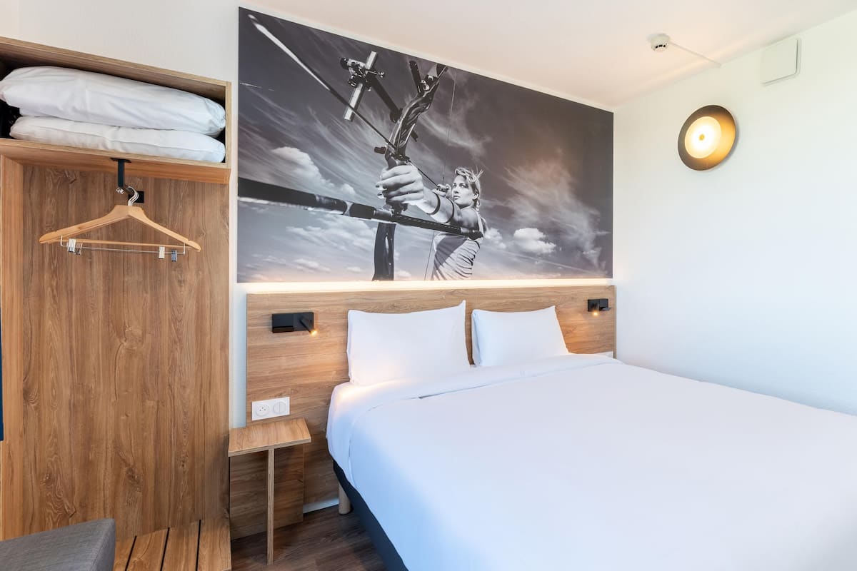 A hotel room with a large bed, a wooden wardrobe, a wall-mounted light, and a painting of a person with a bow.