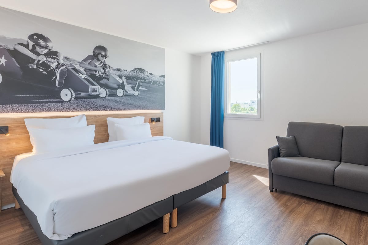 A modern hotel room with a large bed, a grey sofa, a wooden floor, and a wall-mounted artwork.