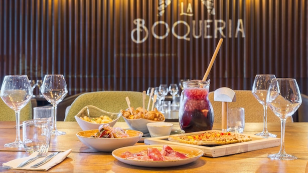 A dining table with various dishes, glasses, and a drink, set against a backdrop with vertical lines and the word 'BOQUERIA' visible. - Campanile PRIME - Lyon Meyzieu Eurexpo Arena Stadium (Meyzieu)