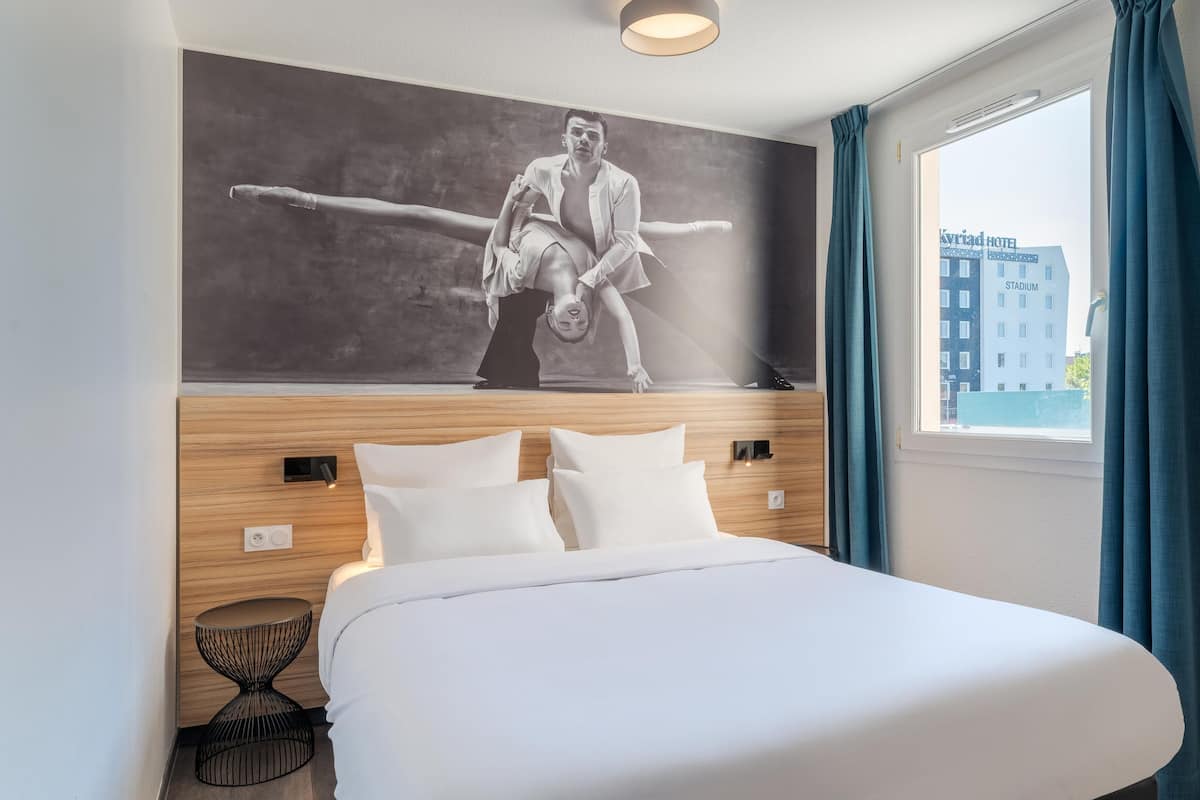 A hotel room with a large bed, a wall-mounted painting, a window with curtains, and a view of a building outside.