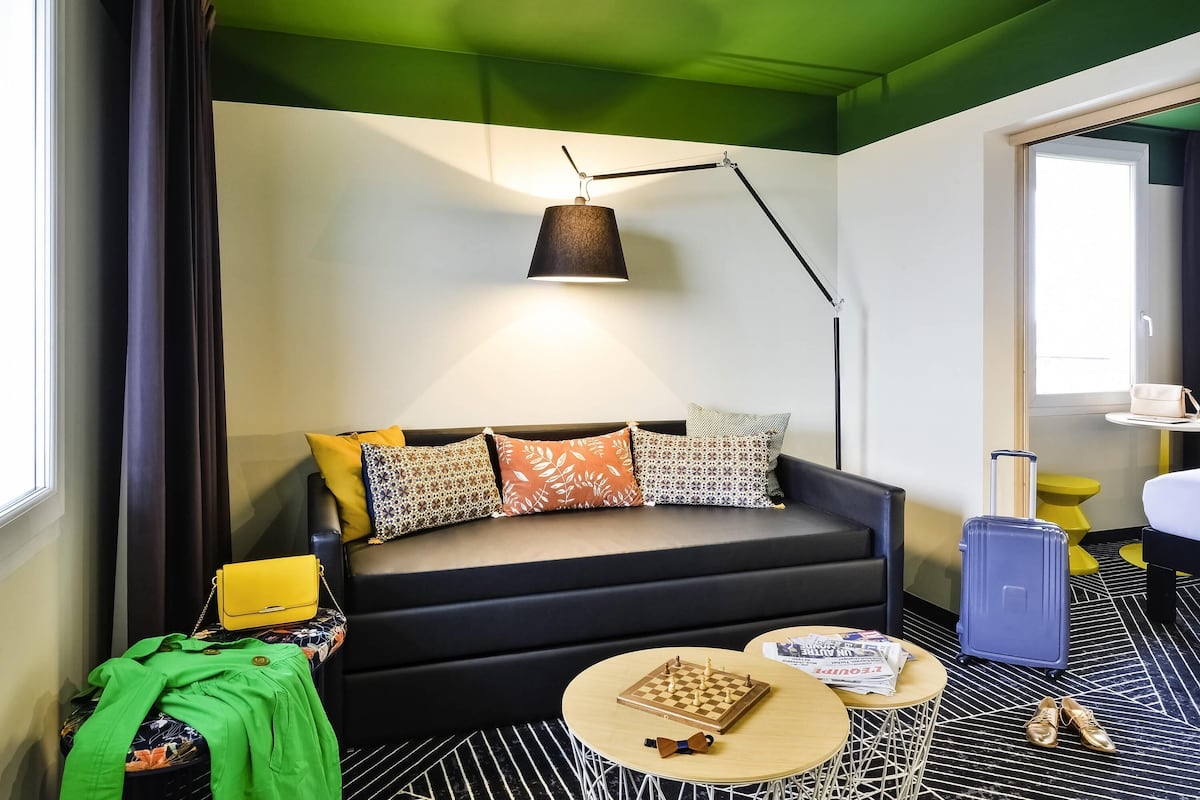 Habitación de hotel con sofá, lámpara, tablero de ajedrez, maleta y un par de zapatos.