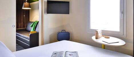 Una habitación de hotel con una cama, una mesita con una revista, una maleta, un televisor en la pared y una ventana con luz natural.