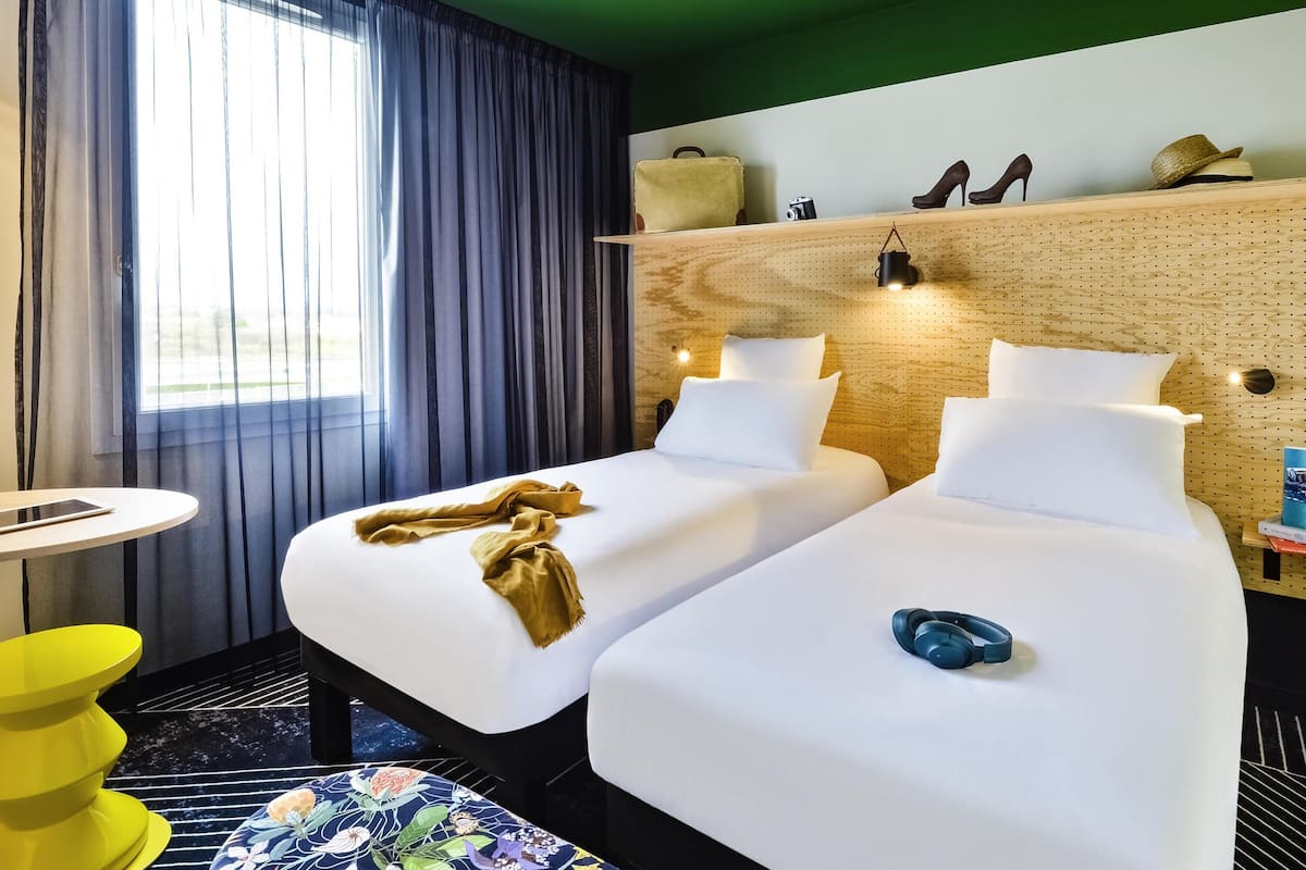 Habitación de hotel con dos camas, una mesita pequeña, una silla amarilla, un ventanal con cortinas y un cabecero de madera con luces.
