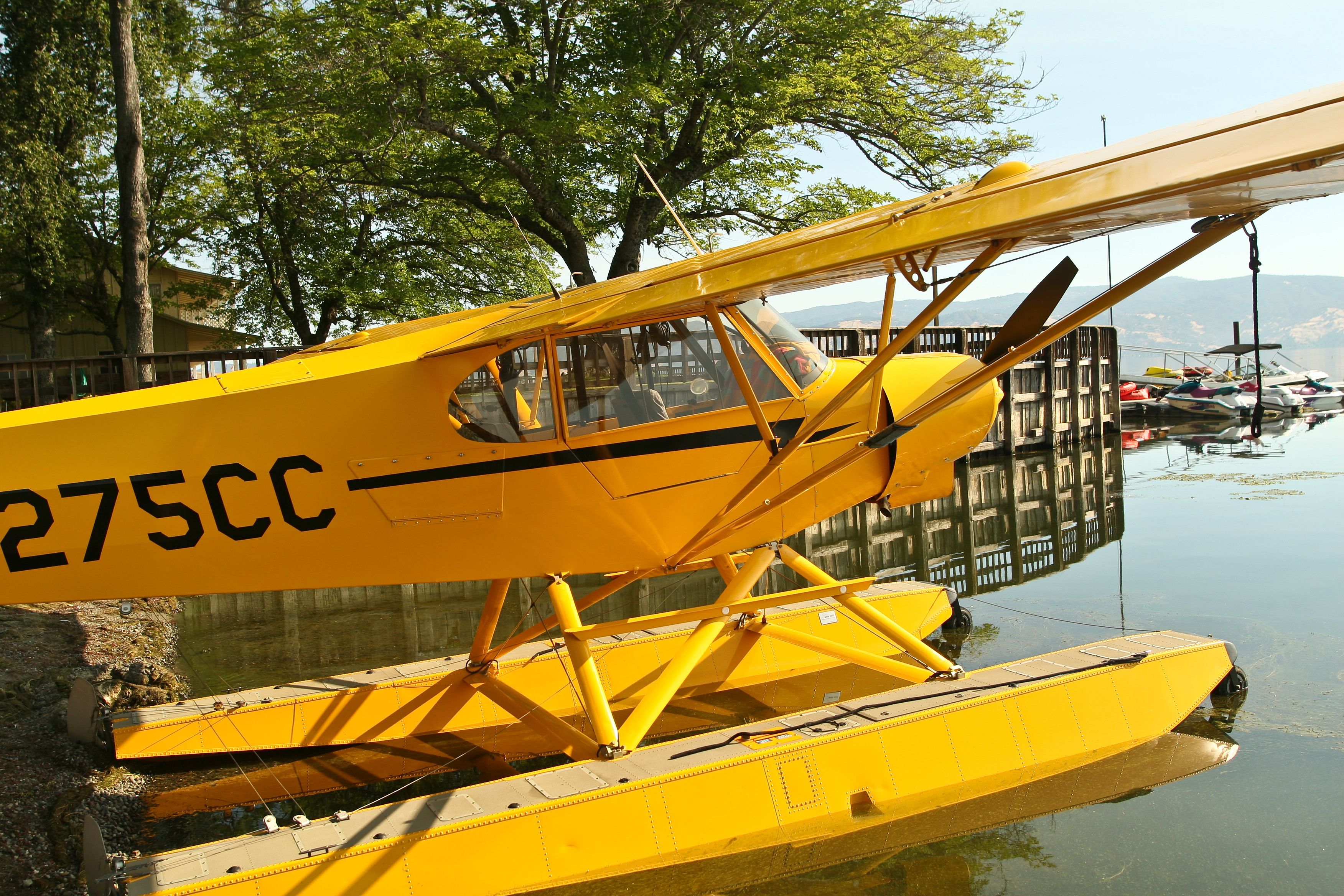 A yellow seaplane with the registration number 275CC parked on a body of water, surrounded by trees and other boats.