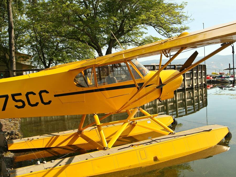 A yellow seaplane with the registration number 275CC parked on a body of water, surrounded by trees and other boats.
