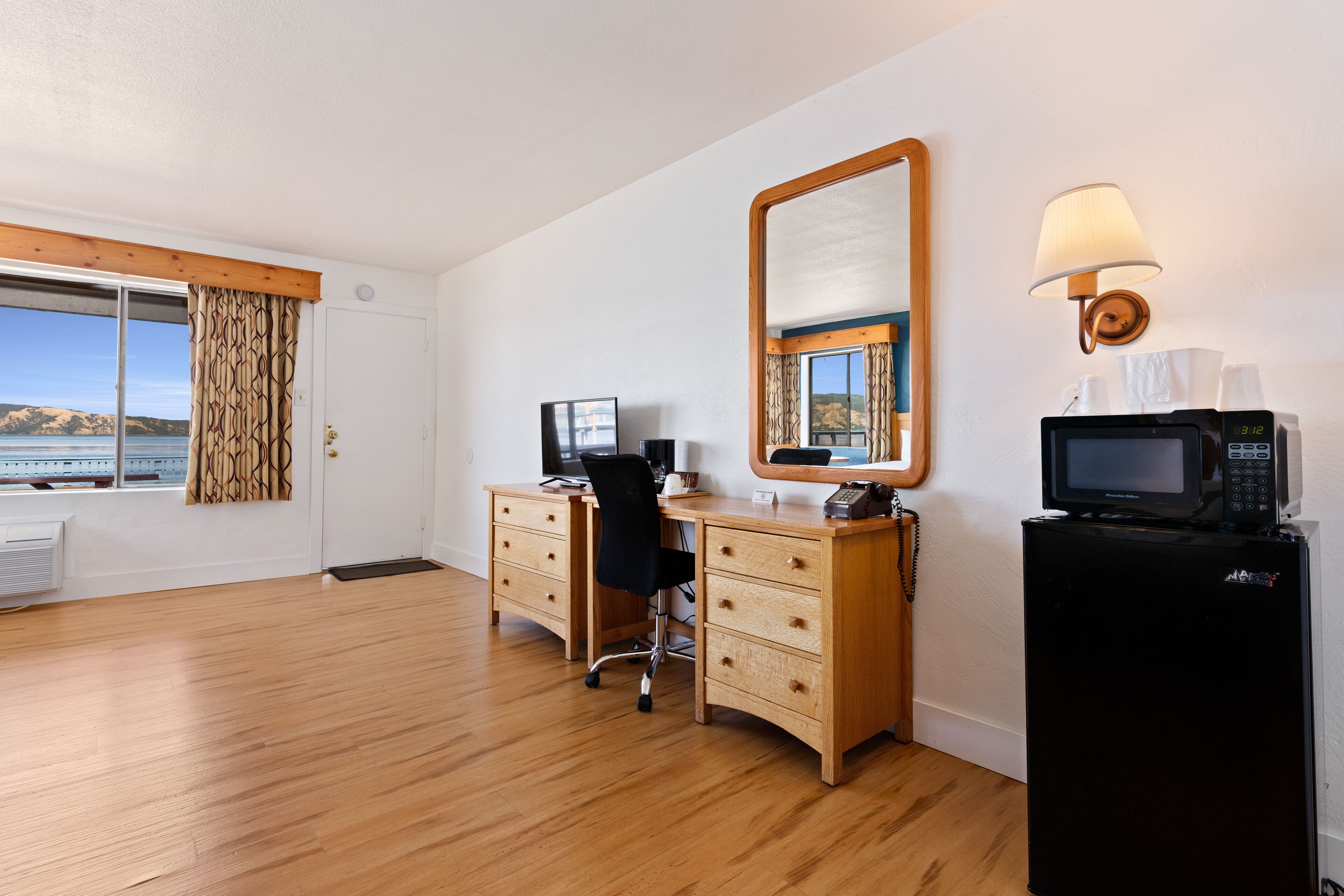 Habitación de hotel con escritorio de madera, computadora, refrigerador negro, microondas, espejo y ventana con vista al mar.