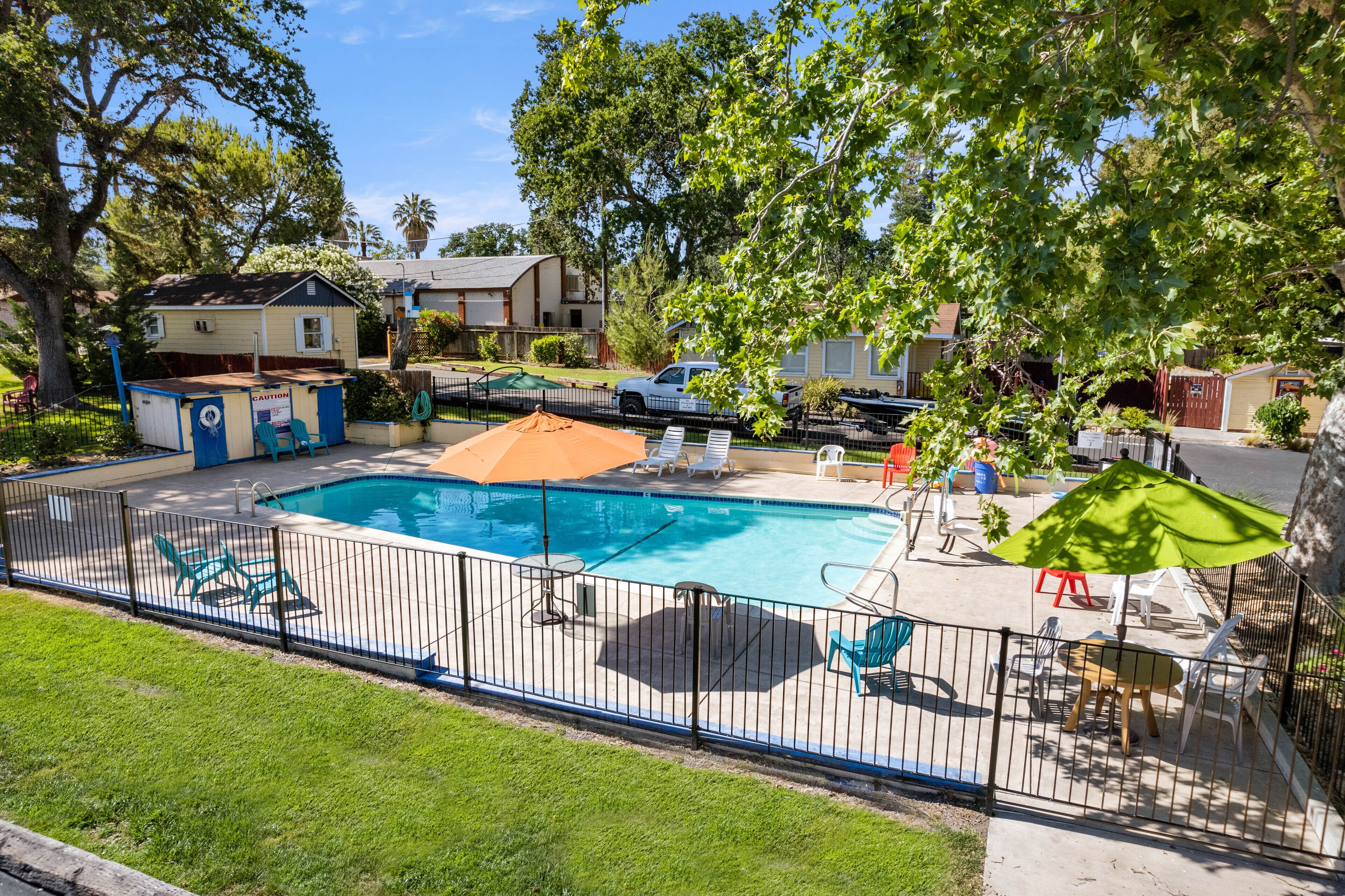 Une piscine rectangulaire entourée d’une terrasse délimitée par une clôture, le tout bordé d’arbres et adossé à un petit bâtiment.