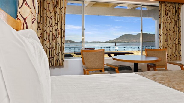 A hotel room with a large bed, two chairs, a table, and a view of the sea and mountains through large windows.