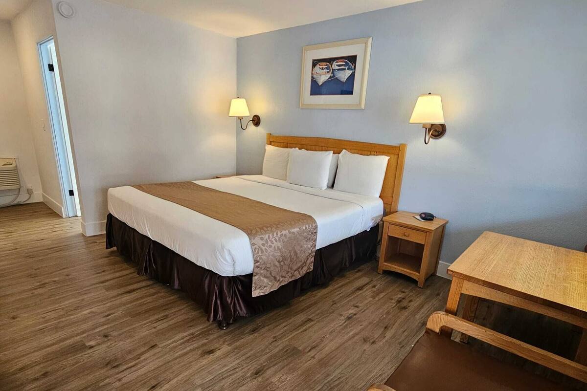 Una habitación de hotel con una cama grande, dos lámparas de noche, una mesita de noche de madera, una pequeña mesa y una puerta que da a otra habitación.